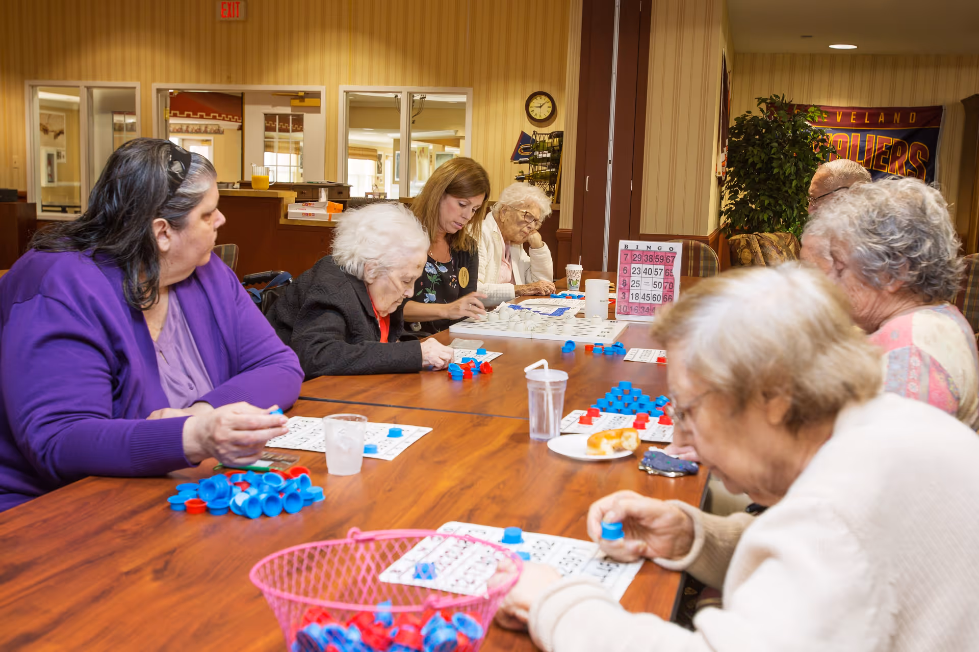 A group of elderly women and a caregiver sitting around a wooden table playing bingo. The table is covered with bingo cards and blue and red bingo chips. The room has beige striped wallpaper, a clock on the wall, and a Cleveland Cavaliers banner in the background.