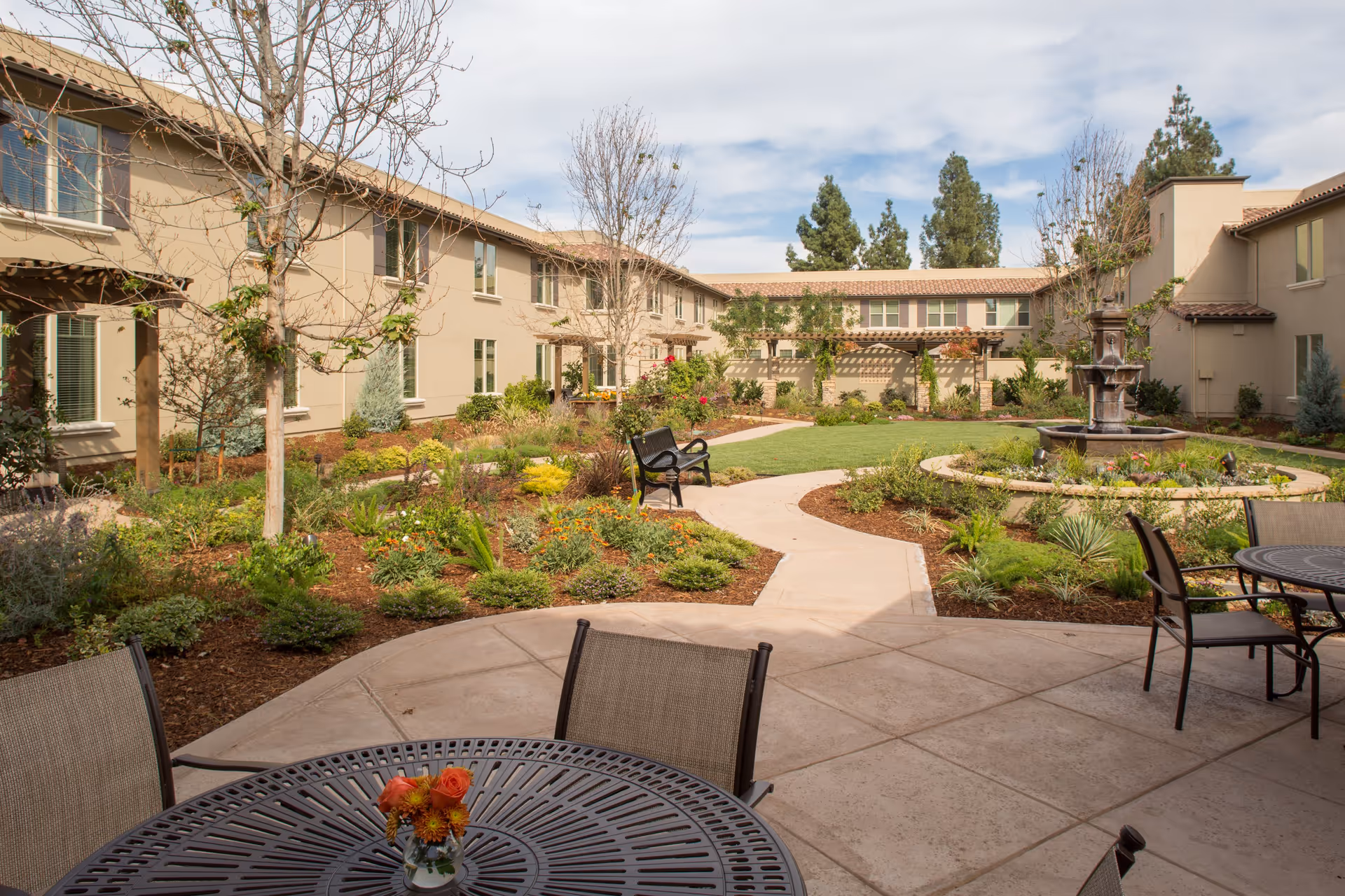 Outdoor courtyard area of a senior living facility with a paved patio featuring round metal tables and chairs, a landscaped garden with various plants and flowers, a central water fountain, and a two-story beige building surrounding the courtyard under a partly cloudy sky.