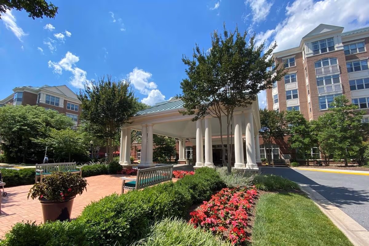 Outdoor view of Maplewood Park Place featuring a covered entrance with white columns, surrounded by well-maintained landscaping including green bushes, red flowers, and trees. The building is a multi-story brick structure under a blue sky with scattered clouds.