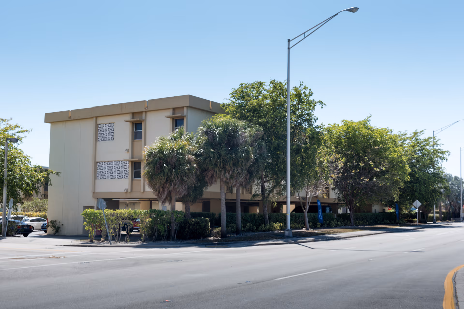 Exterior view of a three-story beige building partially obscured by palm trees and other greenery along a street with a sidewalk and streetlights under a clear blue sky.