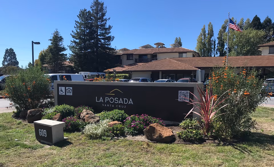 Outdoor view of La Posada Retirement Community sign surrounded by plants and flowers with a building and parked cars in the background under a clear blue sky.