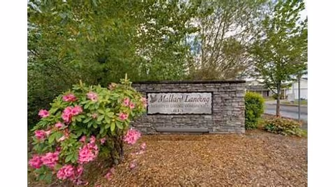 Stone sign for Mallard Landing Assisted Living Community surrounded by greenery and pink flowers, with trees and a building visible in the background.