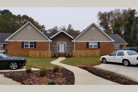 Single-story brick and siding apartment building with a central entrance, curved walkway, small fenced porches, and parked cars in the front.