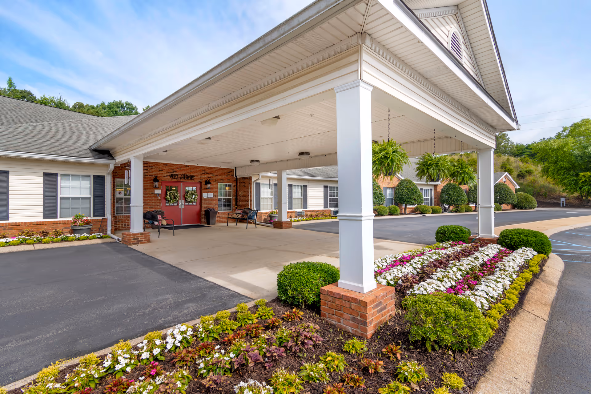 Entrance of Brookdale Hixson senior living facility with a covered drop-off area supported by white columns. The building has red double doors with wreaths, brick and white siding exterior, and black shutters on windows. There are benches near the entrance and a well-maintained flower bed with colorful flowers and green shrubs in front. The sky is clear with some clouds.