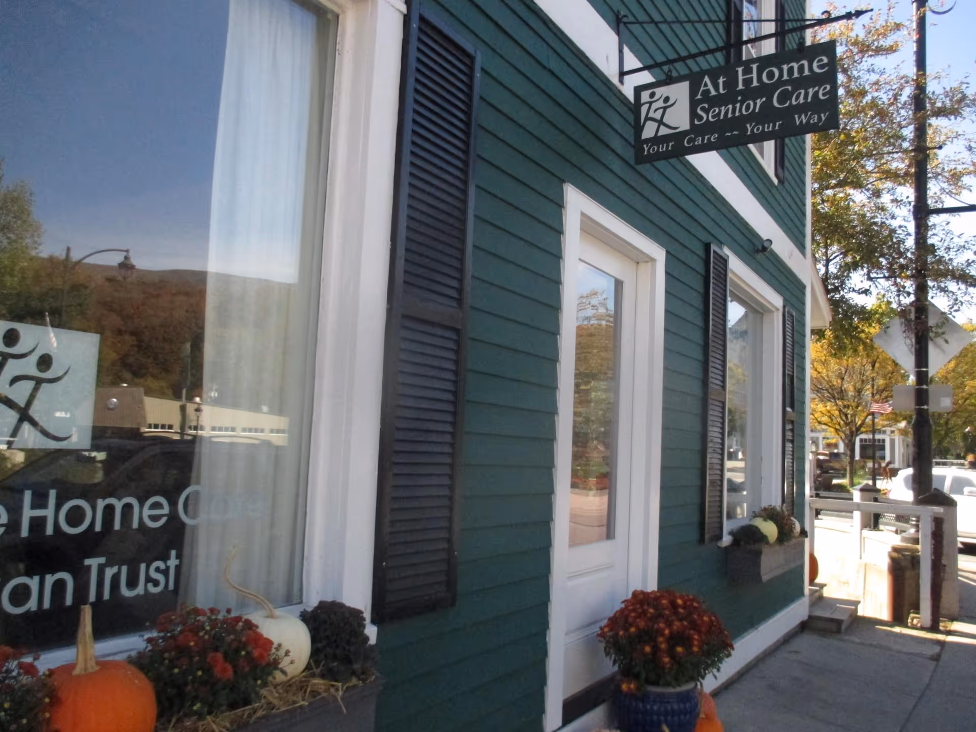 Front exterior of a green 'At Home Senior Care' storefront with a hanging sign, entrance door, window display and pumpkins and potted flowers by the sidewalk.