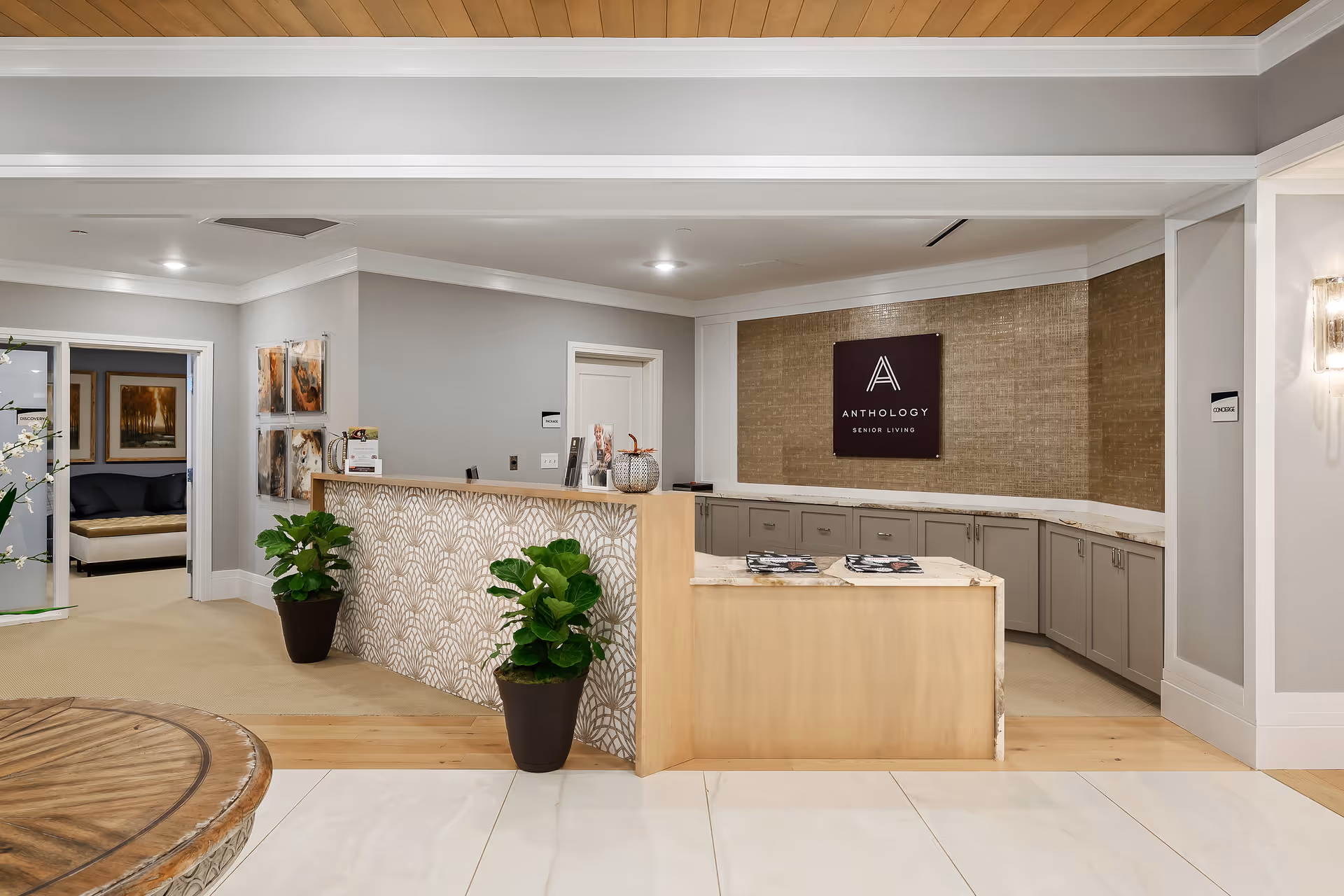 Reception area of a senior living facility with a light wood and patterned front desk, two potted plants, and a sign on the back wall that reads 'Anthology Senior Living'. The space has light-colored walls, recessed lighting, and a seating area visible through an open doorway on the left.
