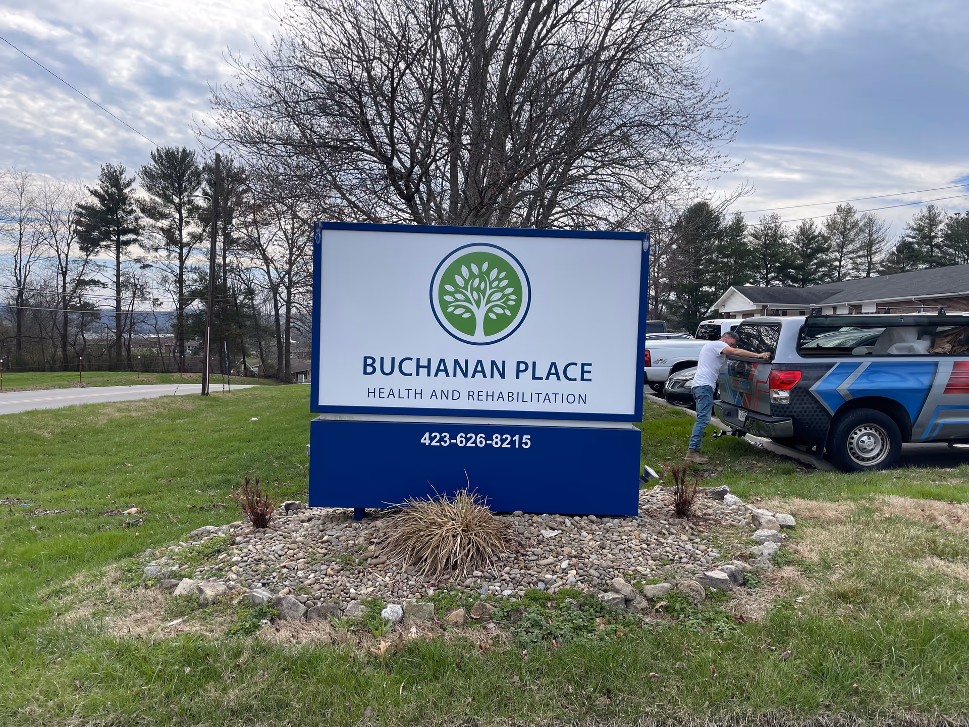Outdoor view of a sign for Buchanan Place Health and Rehabilitation, placed on a grassy area with rocks and plants around its base. In the background, there are trees, a road, and a man leaning into the back of a pickup truck parked near a building.