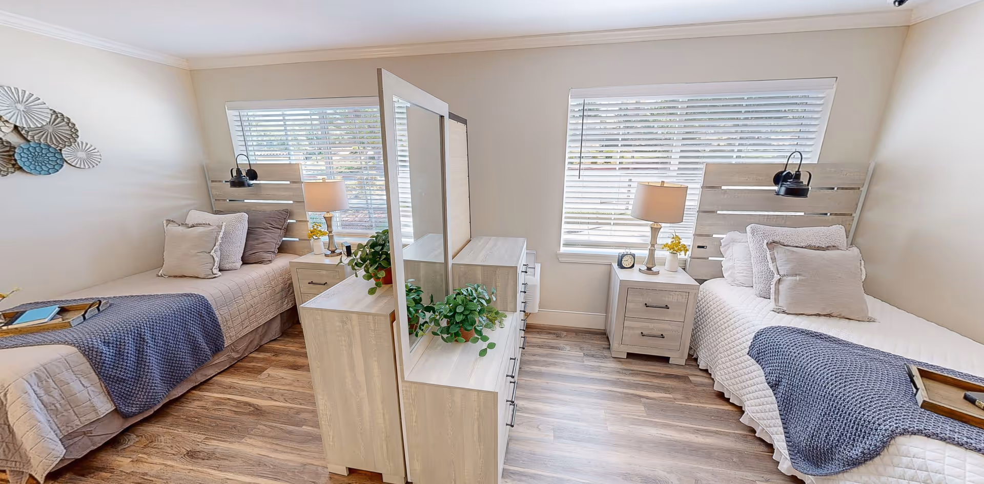 A bright and clean bedroom with two single beds separated by a wooden dresser with a mirror. Each bed has a headboard, pillows, a bedside table with a lamp, and a window with blinds behind it. The room has wooden flooring and neutral-colored walls, decorated with a wall art piece on the left side.