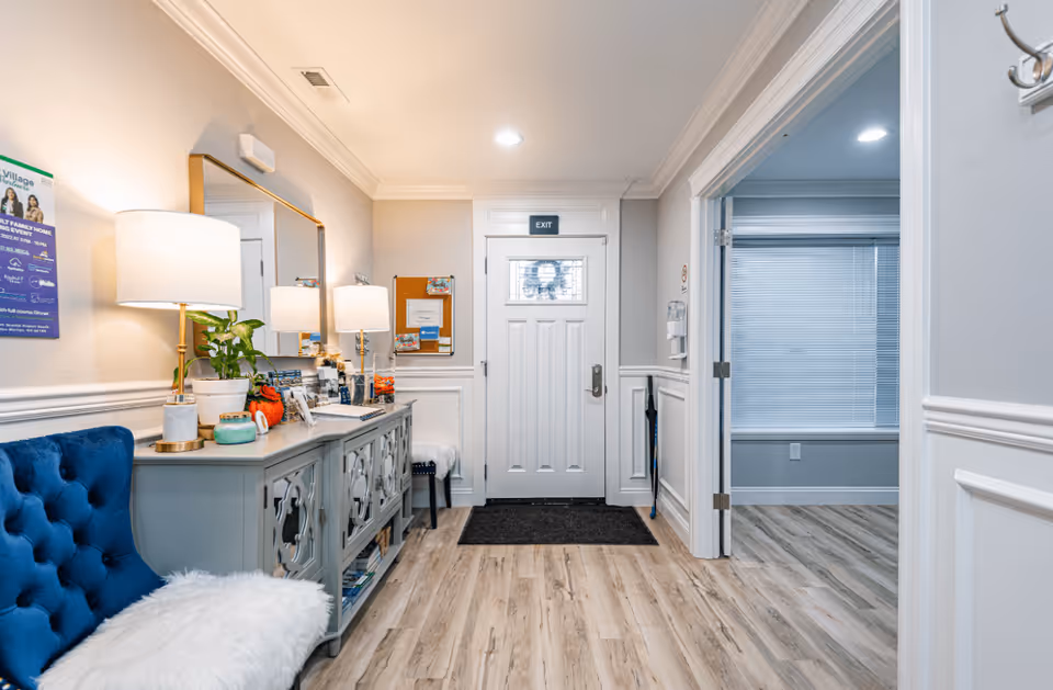 A well-lit entryway of an adult family home with a white front door marked with an exit sign above it. To the left, there is a gray decorative console table with a lamp, plants, and various items on top. A large mirror hangs above the table. A blue cushioned bench with a white furry throw is positioned against the left wall. The floor is light wood, and there is a doorway on the right leading to another room with a window covered by blinds.
