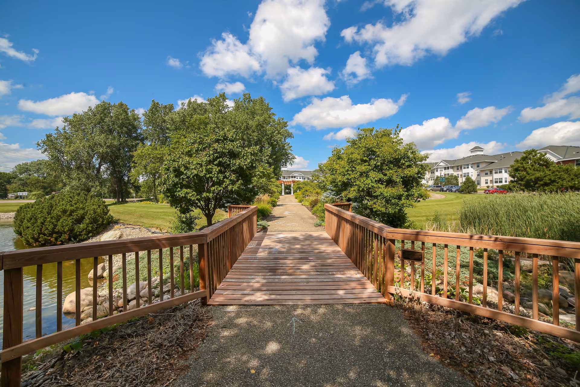 A wooden footbridge over a small pond leading to a paved pathway surrounded by green trees and grass, with a large residential building in the background under a blue sky with scattered clouds.