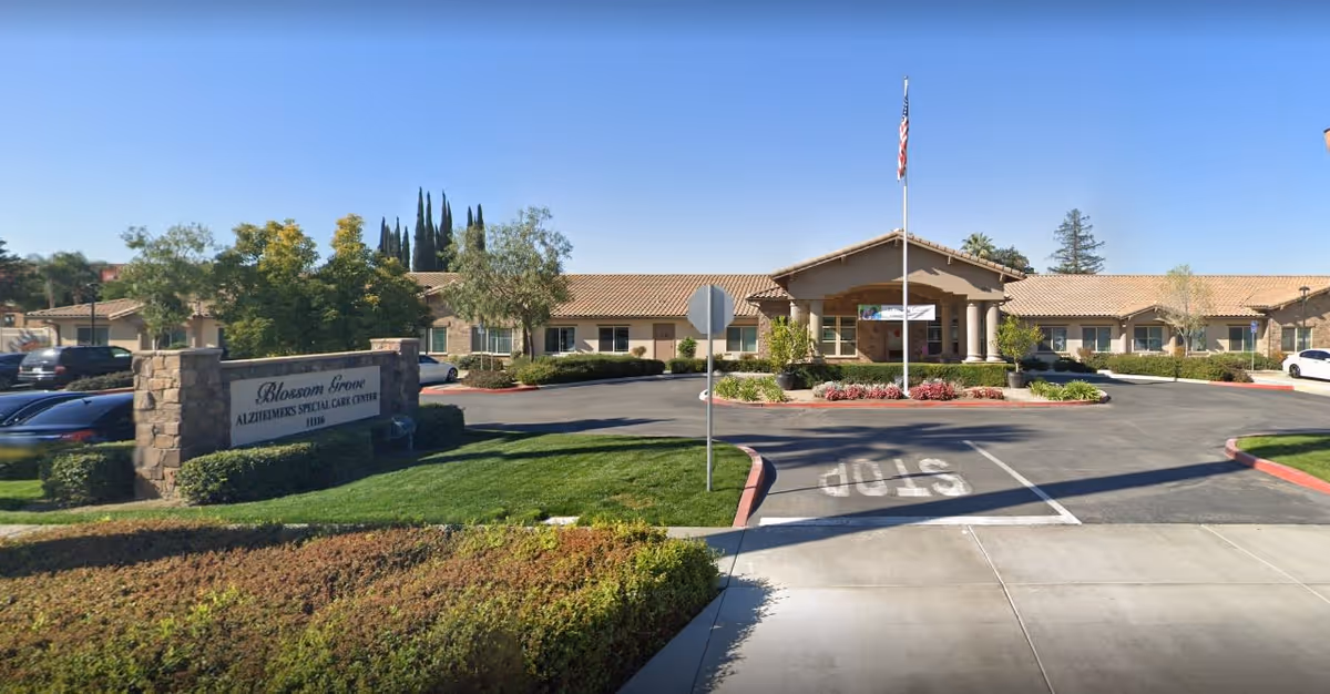 Front exterior view of Blossom Grove Alzheimer's Special Care Center building with a driveway, landscaped greenery, and an American flag on a flagpole in front.