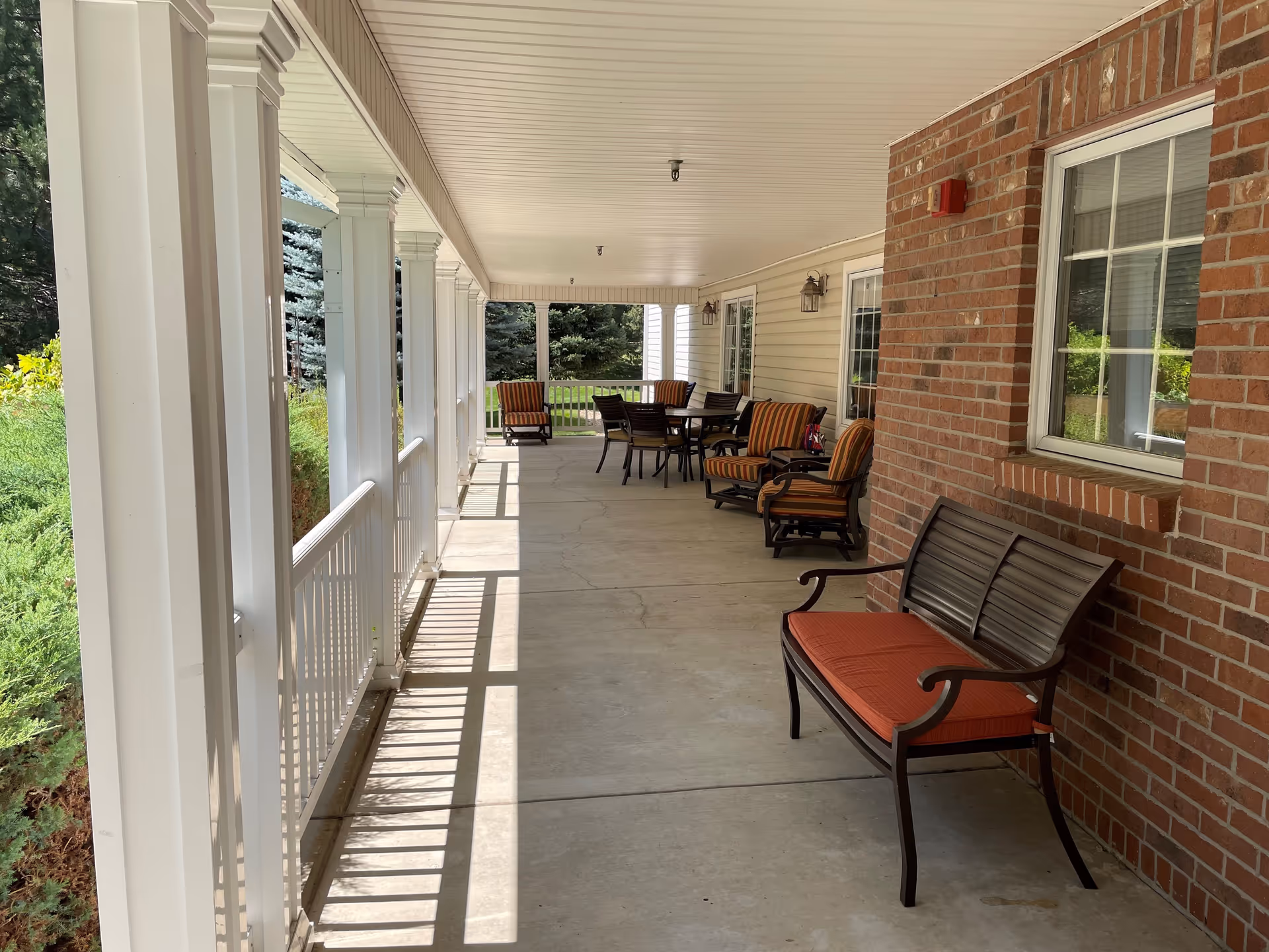 Covered outdoor patio area with white pillars and railing on the left side, and a brick wall with windows on the right side. The patio has several cushioned chairs and a table with chairs arranged for seating. Greenery and trees are visible beyond the railing.