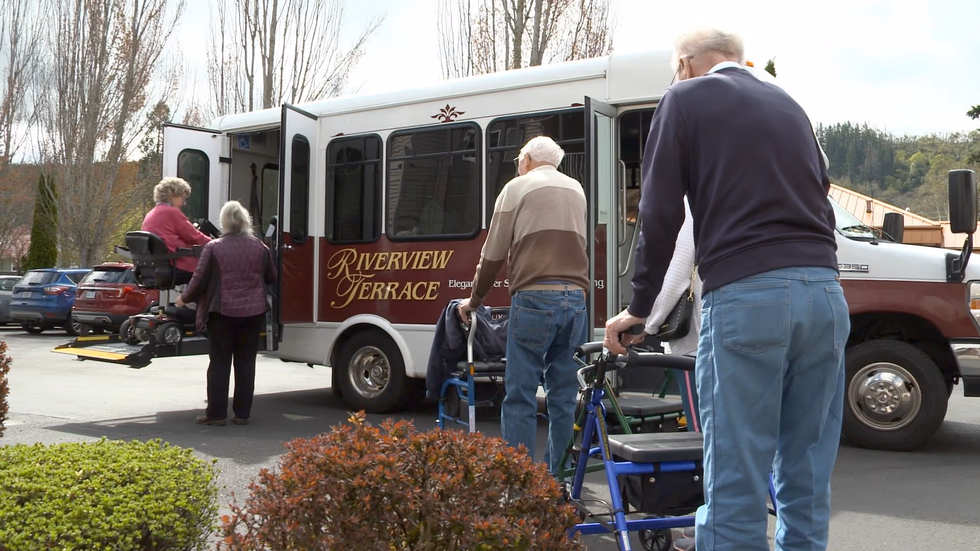 Several elderly individuals using walkers and a mobility scooter boarding a Riverview Terrace shuttle bus in an outdoor parking area with trees and bushes nearby.