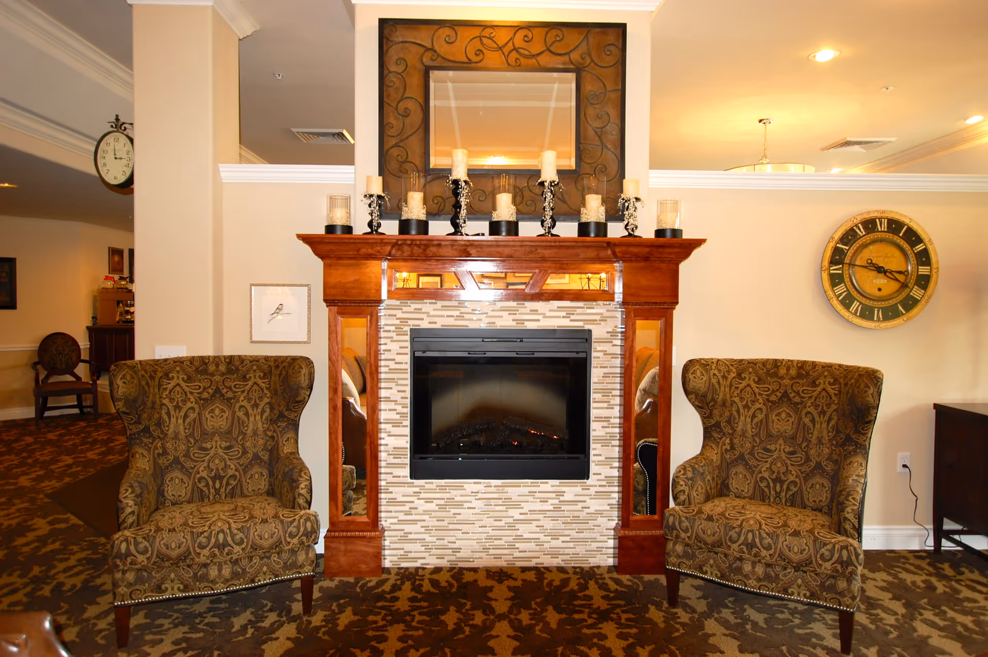 A cozy living room area featuring a decorative fireplace with a wooden mantel and tiled surround. On the mantel are several candles in glass holders. Above the mantel hangs a large ornate mirror. Two patterned wingback chairs are positioned on either side of the fireplace. The room has warm lighting, a patterned carpet, and a large round wall clock on the right wall.