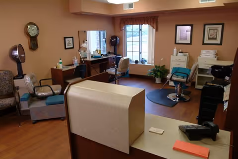 Interior view of a salon area in a senior living facility with hair styling chairs, hair dryers, a reception desk, and framed pictures on the walls. The room has wooden flooring and a window with a valance allowing natural light in.