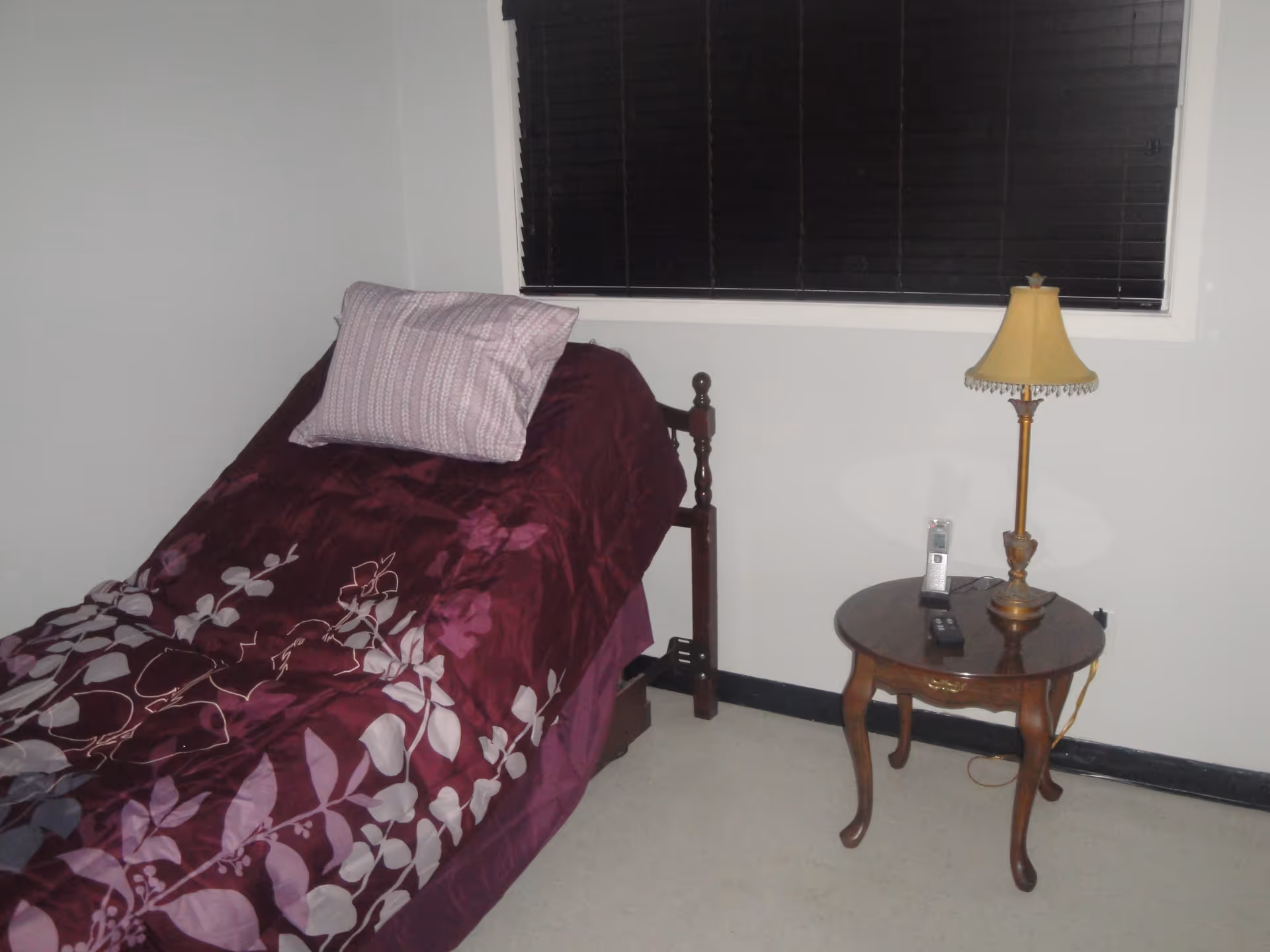 A small bedroom with a single bed covered in a dark purple floral comforter and a light purple pillow. Next to the bed is a wooden side table with a vintage-style lamp, a cordless phone, and a remote control. The window behind the bed has closed dark blinds.