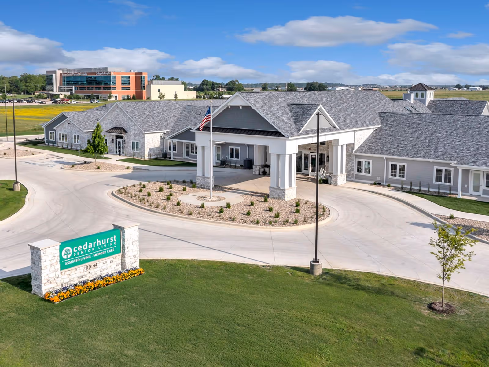 Exterior view of Cedarhurst Senior Living of Newburgh, showing a large single-story building with a covered entrance, a circular driveway, and a flagpole with an American flag. The building has gray siding and a gray shingled roof. In front of the building is a green lawn with a stone sign displaying the facility name and services offered, including assisted living and memory care. The background includes a clear blue sky with some clouds and a distant building labeled 'Orthopedic Hospital.'