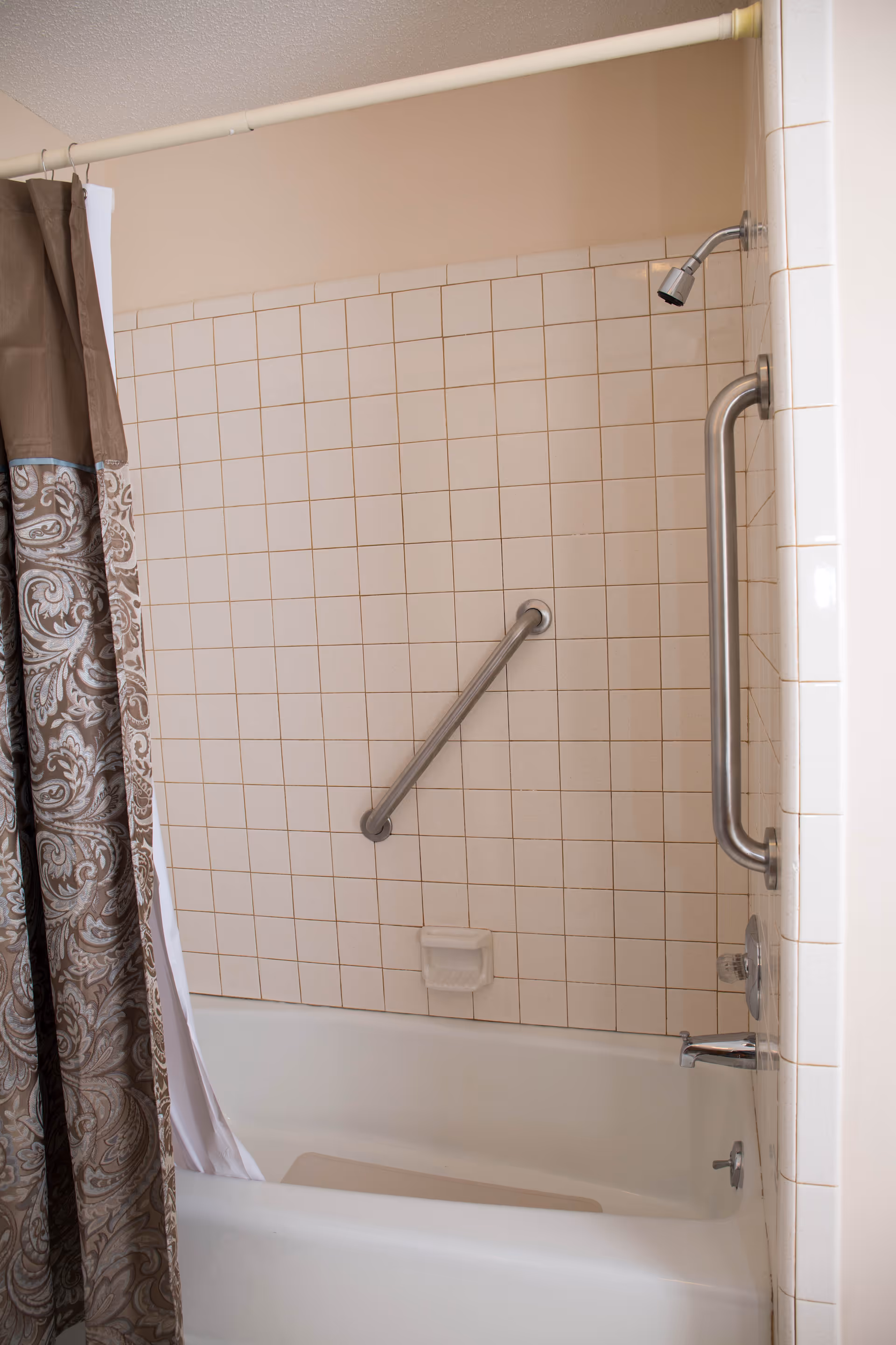 Bathtub and tiled shower area with metal grab bars, a showerhead, and a patterned shower curtain.