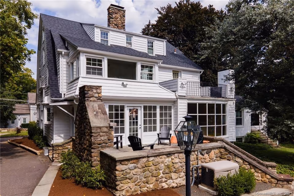 Exterior view of a large white multi-story house with a stone chimney and stone wall patio area. The house has multiple windows, a balcony, and outdoor seating. Trees and greenery surround the building under a partly cloudy sky.
