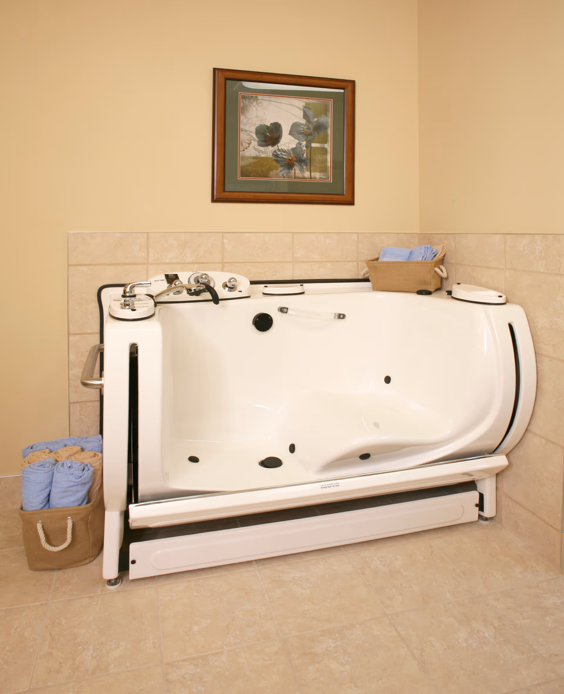 A white walk-in bathtub with safety handles and controls, placed against a beige tiled wall. There are two baskets with rolled towels, one on the floor and one on the tub edge. A framed floral artwork hangs on the wall above the tub.