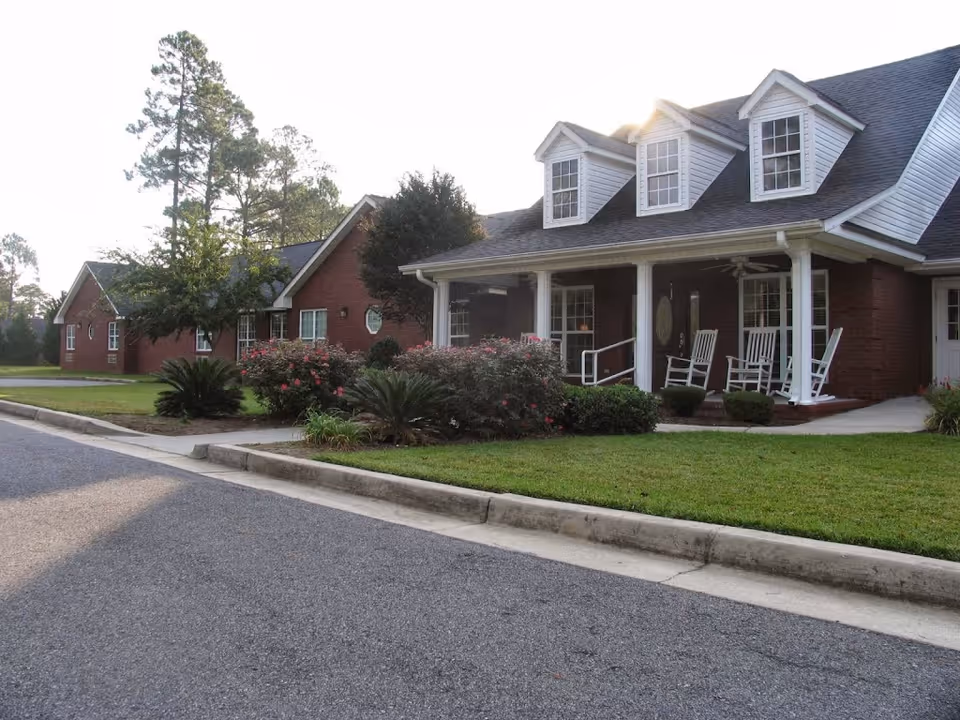 Front exterior of a single-story brick assisted living building with a covered porch, rocking chairs, dormer windows, and landscaped lawn.