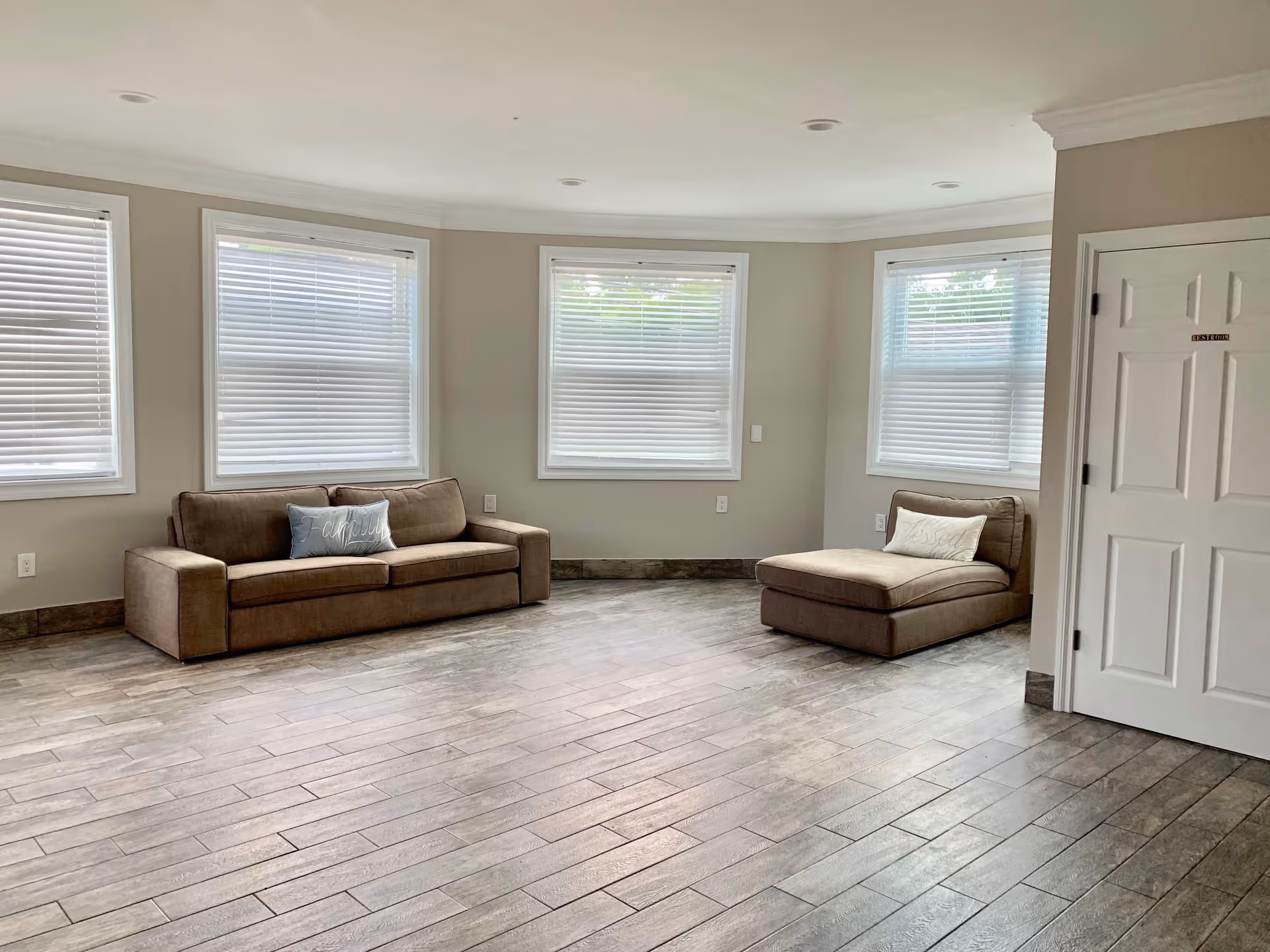 Bright, sparsely furnished living room with a sofa and chaise lounge, multiple windows with blinds, and wood-look tile flooring.