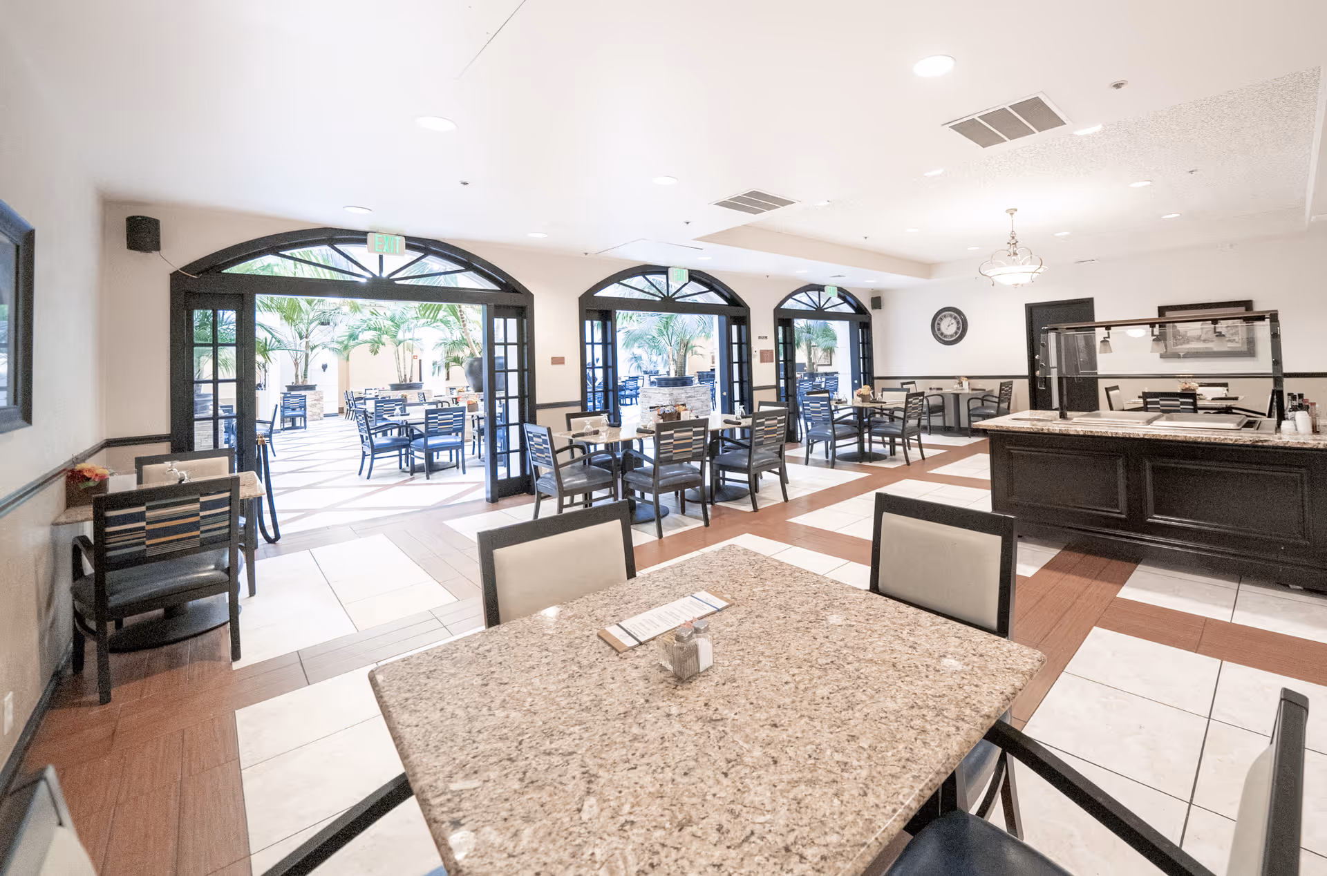 A bright dining area in a senior living facility with multiple tables and chairs. The room features large arched glass doors that open to an outdoor patio with additional seating and palm plants. The interior has a tiled floor with a mix of white and brown tiles, and a serving counter is visible on the right side.