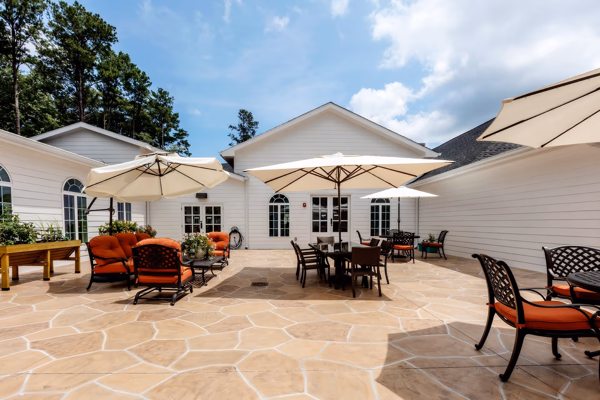 Outdoor patio area at The Mann House Cumming with several seating arrangements including cushioned chairs and tables under large beige umbrellas, surrounded by white buildings and trees under a partly cloudy sky.