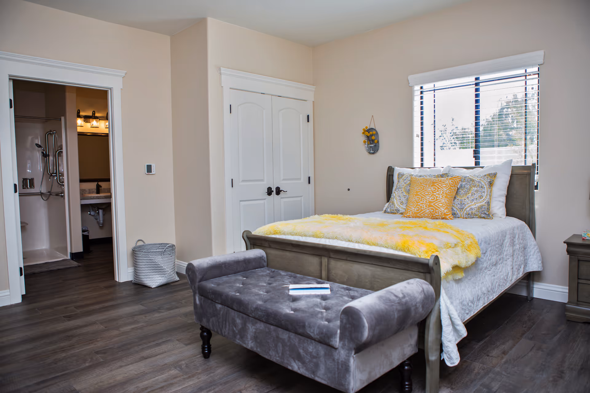 Well-lit bedroom with a bed topped by yellow pillows and throw, a tufted bench at the foot, closet doors, a window, and an open doorway leading to a bathroom.