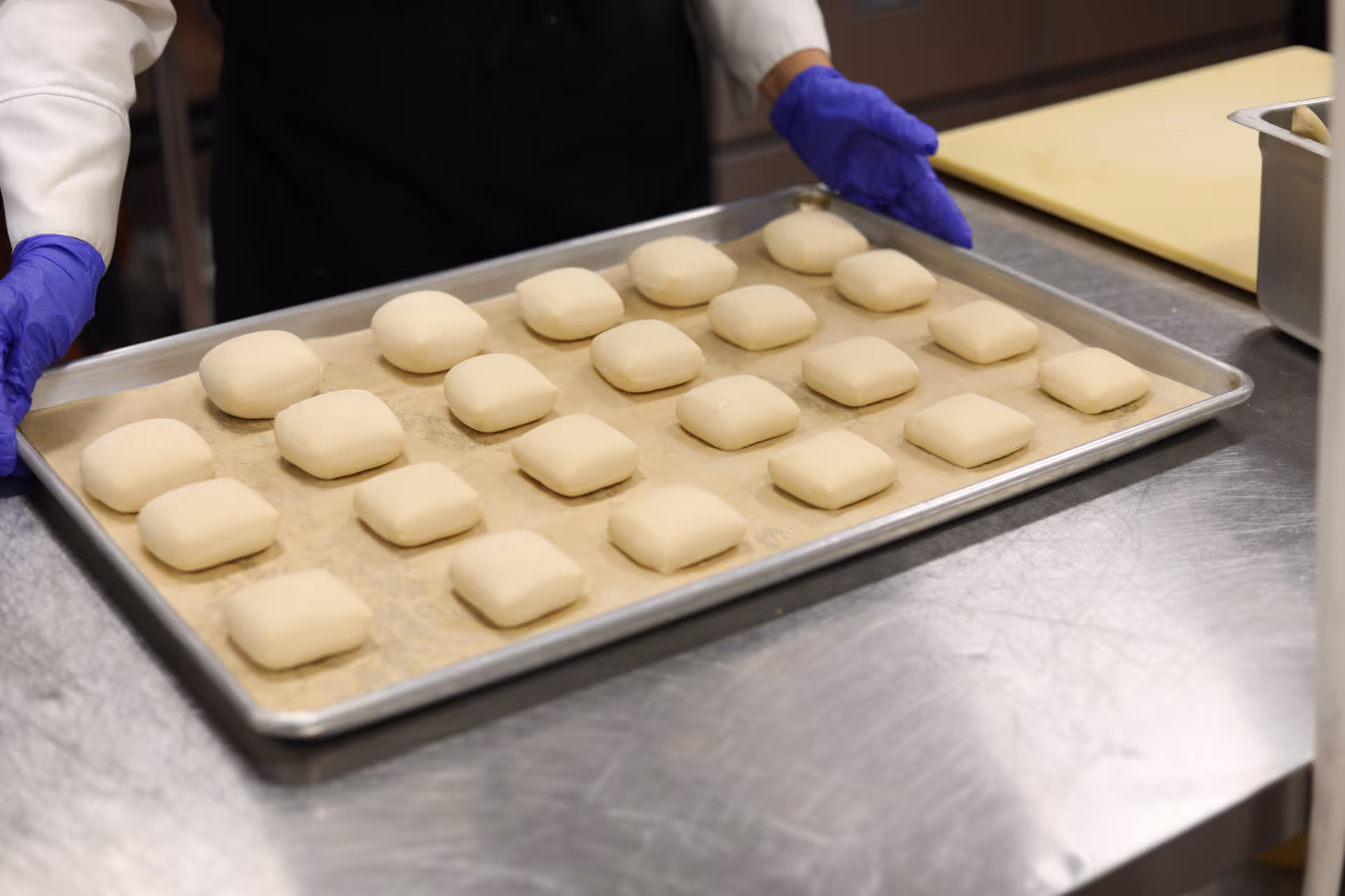 A person wearing purple gloves and a white chef coat holds a baking tray lined with parchment paper, containing rows of small, square dough pieces ready for baking in a commercial kitchen setting.