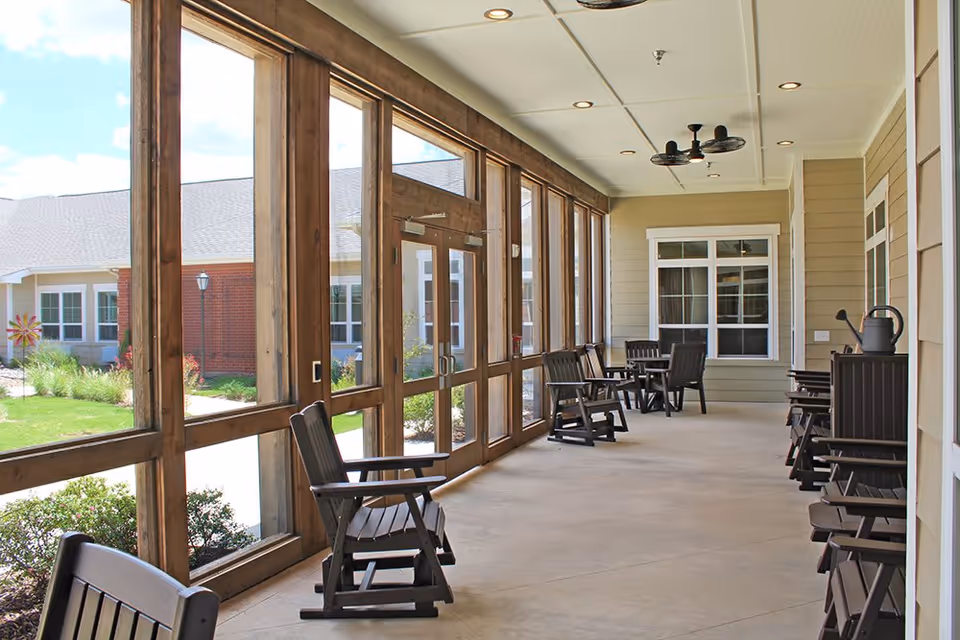 A covered outdoor patio area at Lyndale Abilene Memory Care with multiple wooden rocking chairs and tables. The patio is enclosed with large windows and doors, allowing a view of the garden and building exterior. The ceiling has recessed lighting and a ceiling fan.