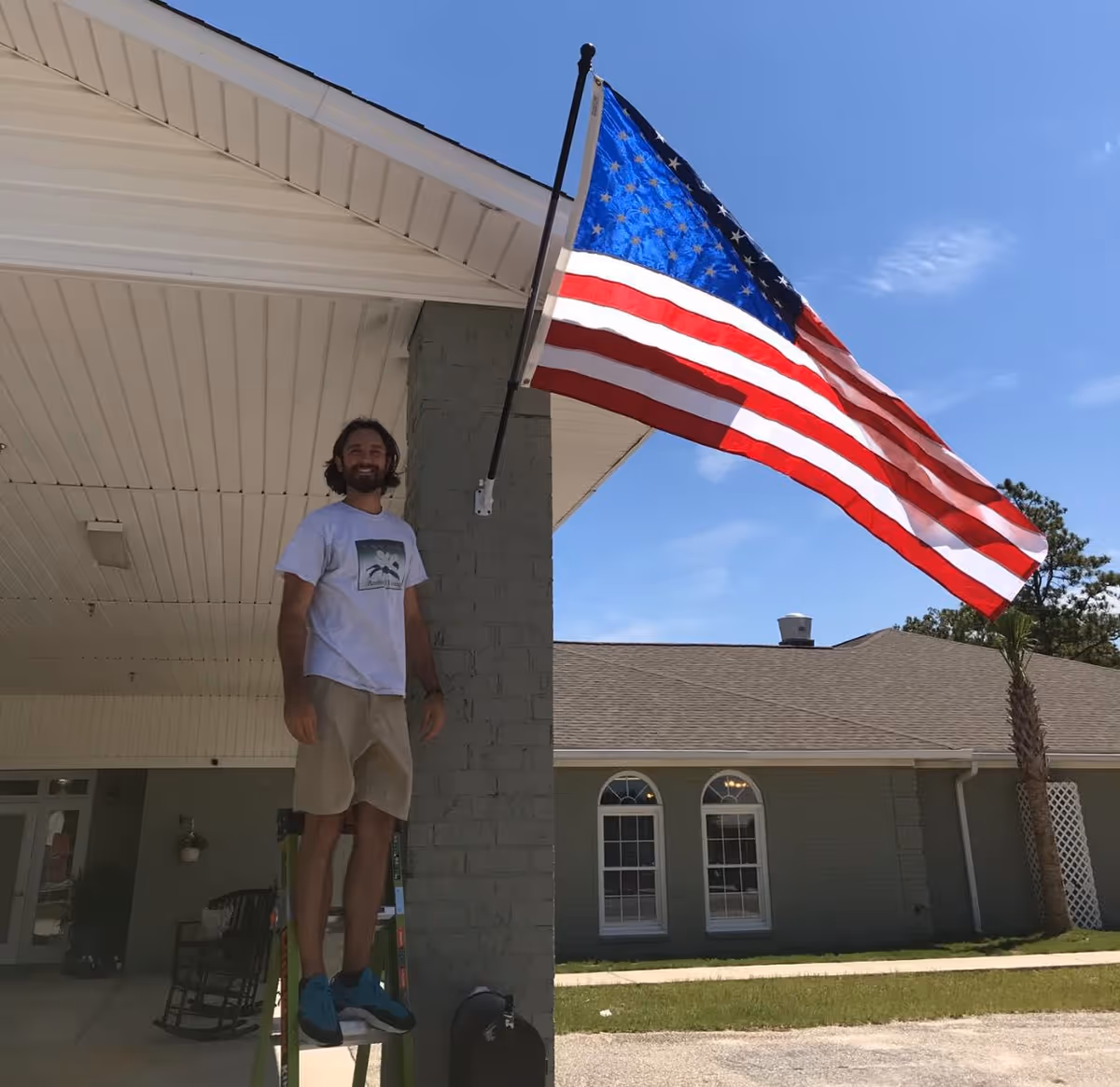 A man standing on a ladder next to a gray brick pillar under a white roof overhang, holding an American flag mounted on the pillar. In the background, there is a building with arched windows, a palm tree, and a clear blue sky.