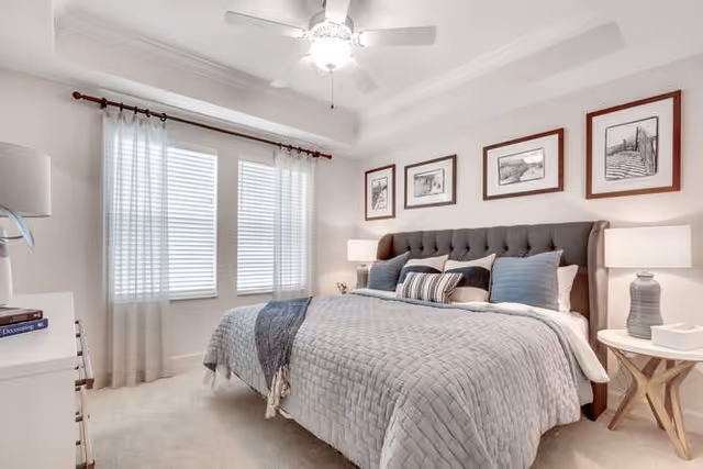 Light-filled bedroom with a large upholstered bed, bedside tables and lamps, framed artwork, and a ceiling fan.