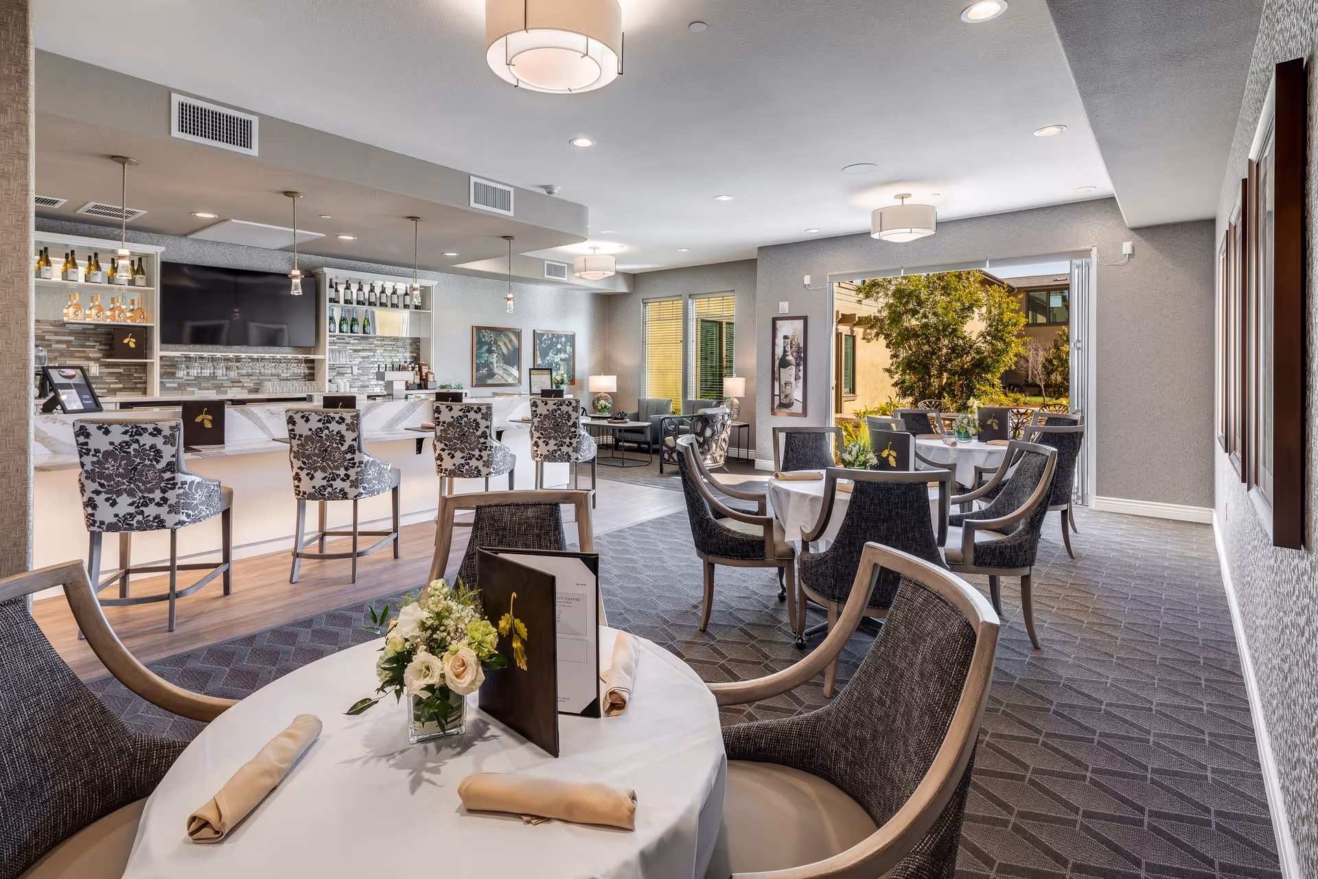 Elegant dining and bar area with round tables, upholstered chairs, patterned bar stools at a counter, and an open doorway to a patio.