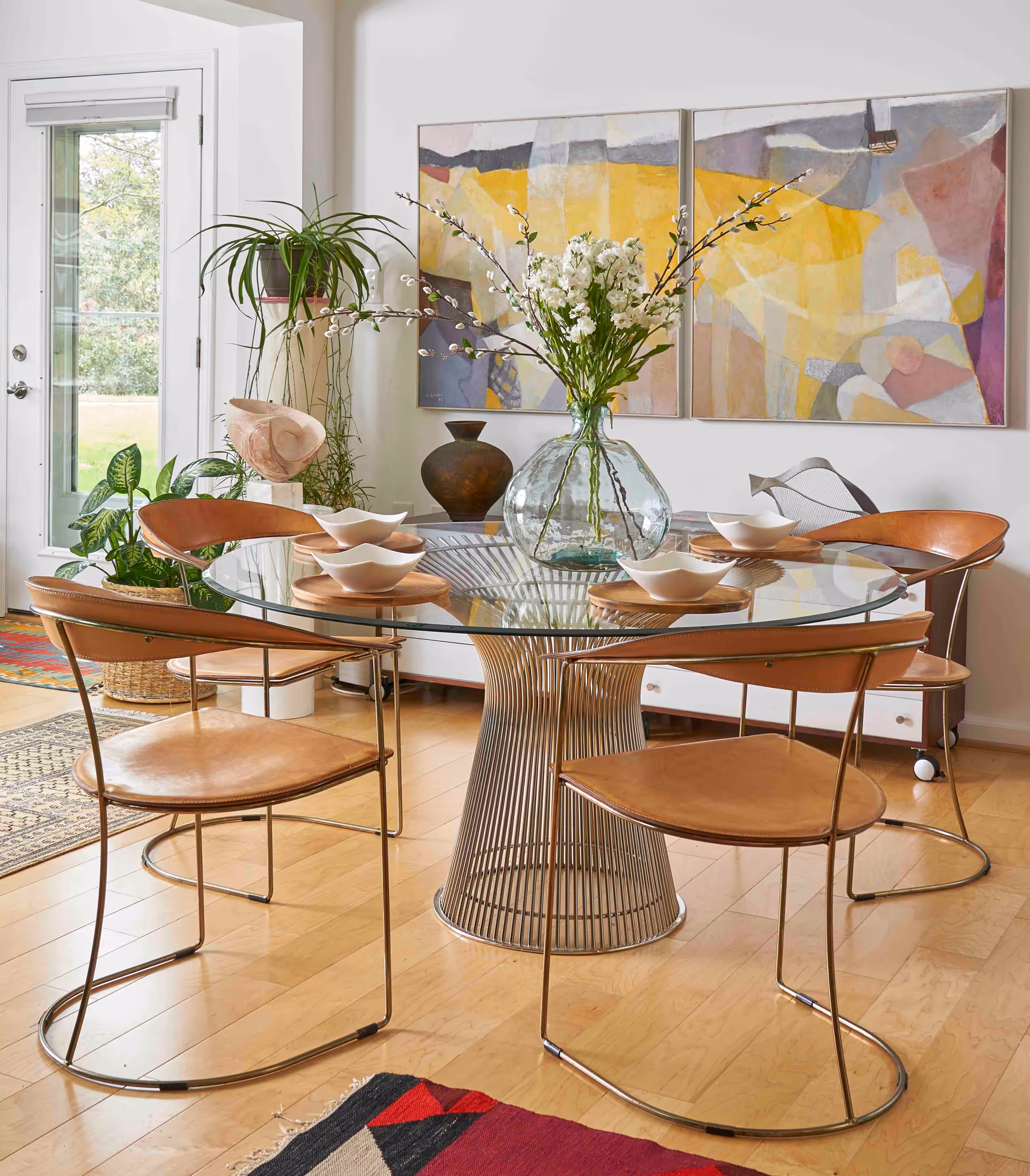 Bright dining room with a round glass table surrounded by leather chairs, a large vase of flowers, plants, and abstract wall art.