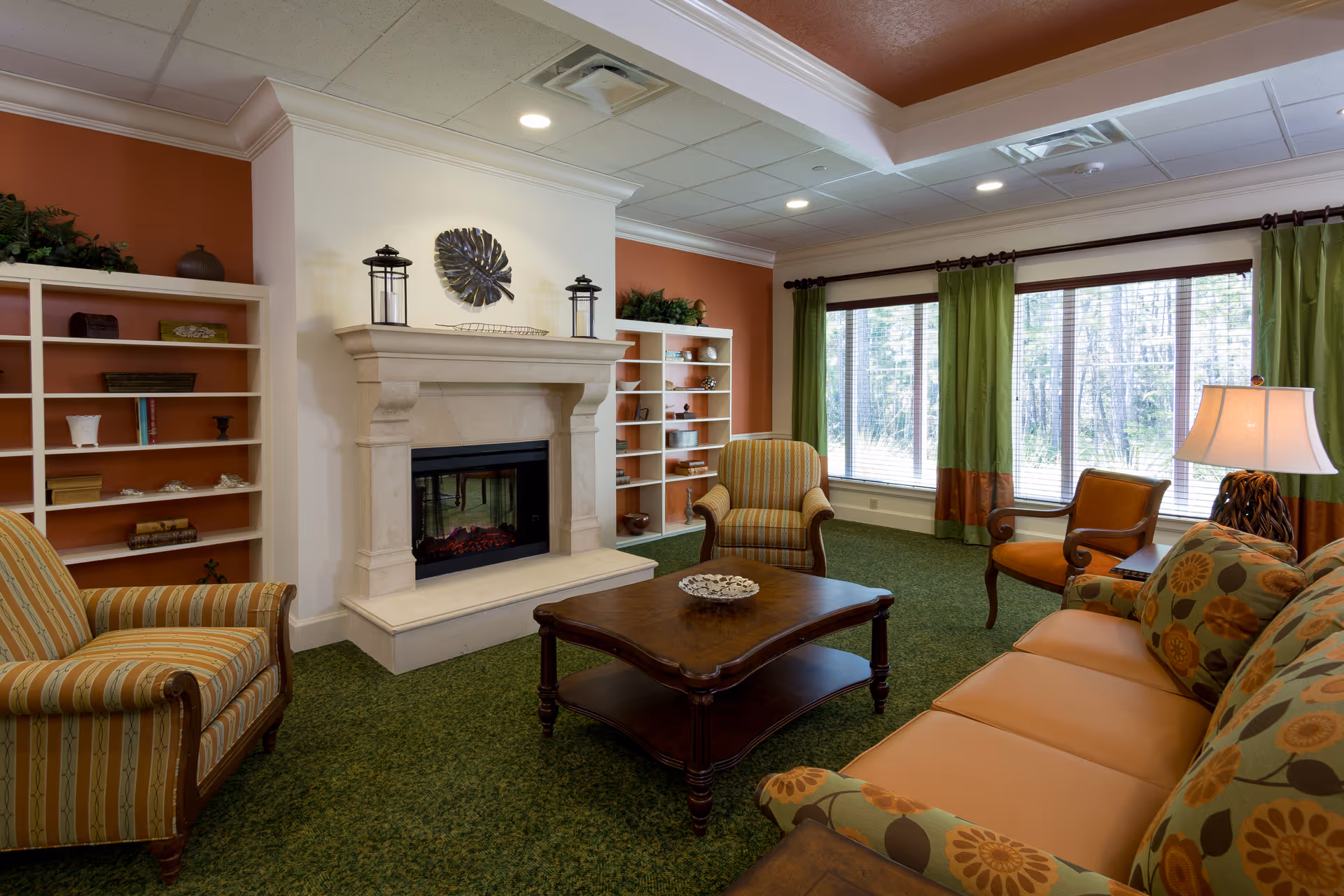 A cozy living room with a green carpet, a beige stone fireplace in the center flanked by white built-in shelves with decorative items. The room has a large window with green and brown curtains letting in natural light. There are multiple seating options including a floral patterned sofa, a striped armchair, and two wooden chairs with cushions. A wooden coffee table with a decorative bowl sits in the middle of the seating area, and a table lamp is on a side table next to the sofa.