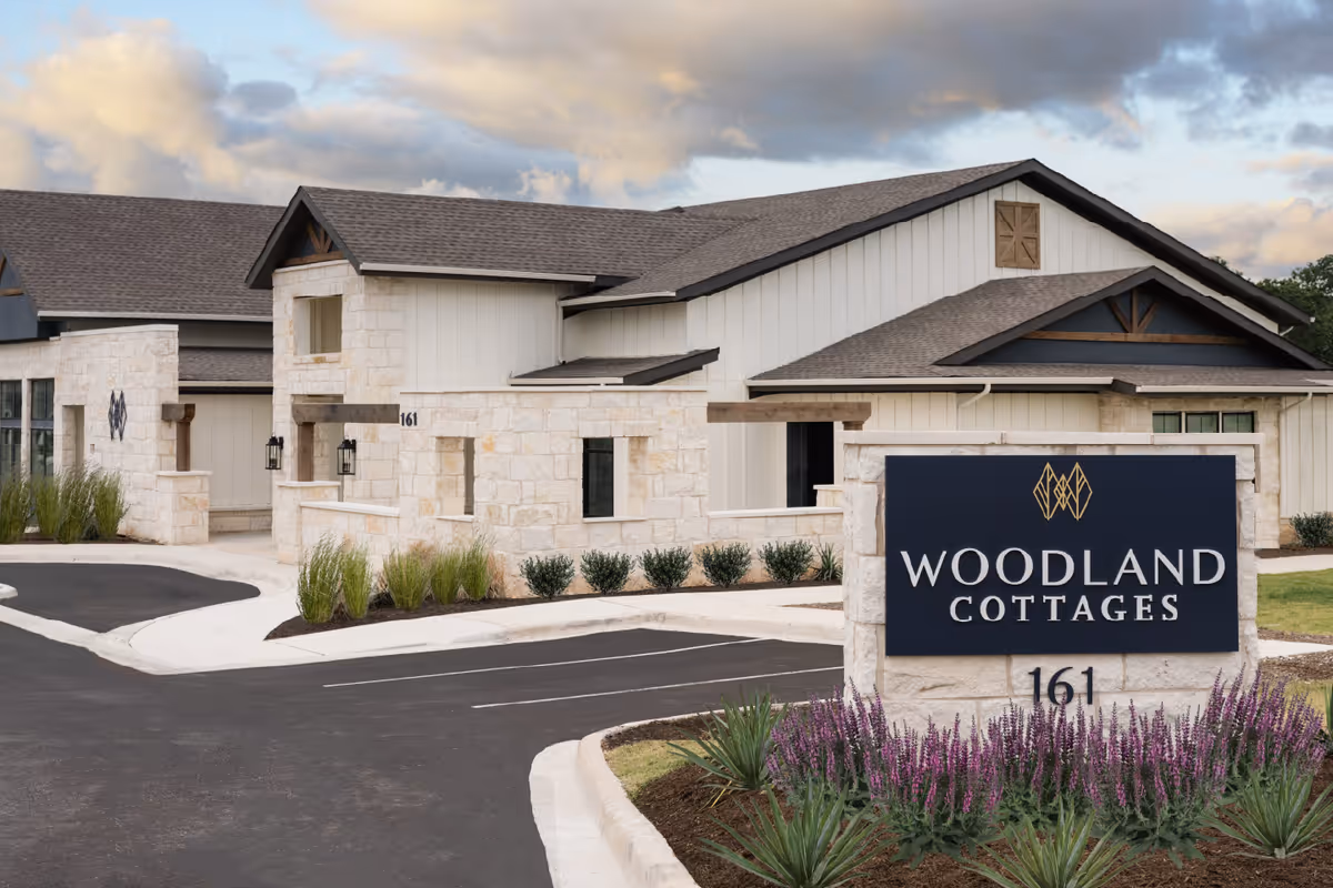Exterior view of Woodland Cottages Senior Living Fredericksburg building with stone and wood siding, a dark shingled roof, and a landscaped parking area. A large sign in front reads 'Woodland Cottages 161' with decorative plants around it.
