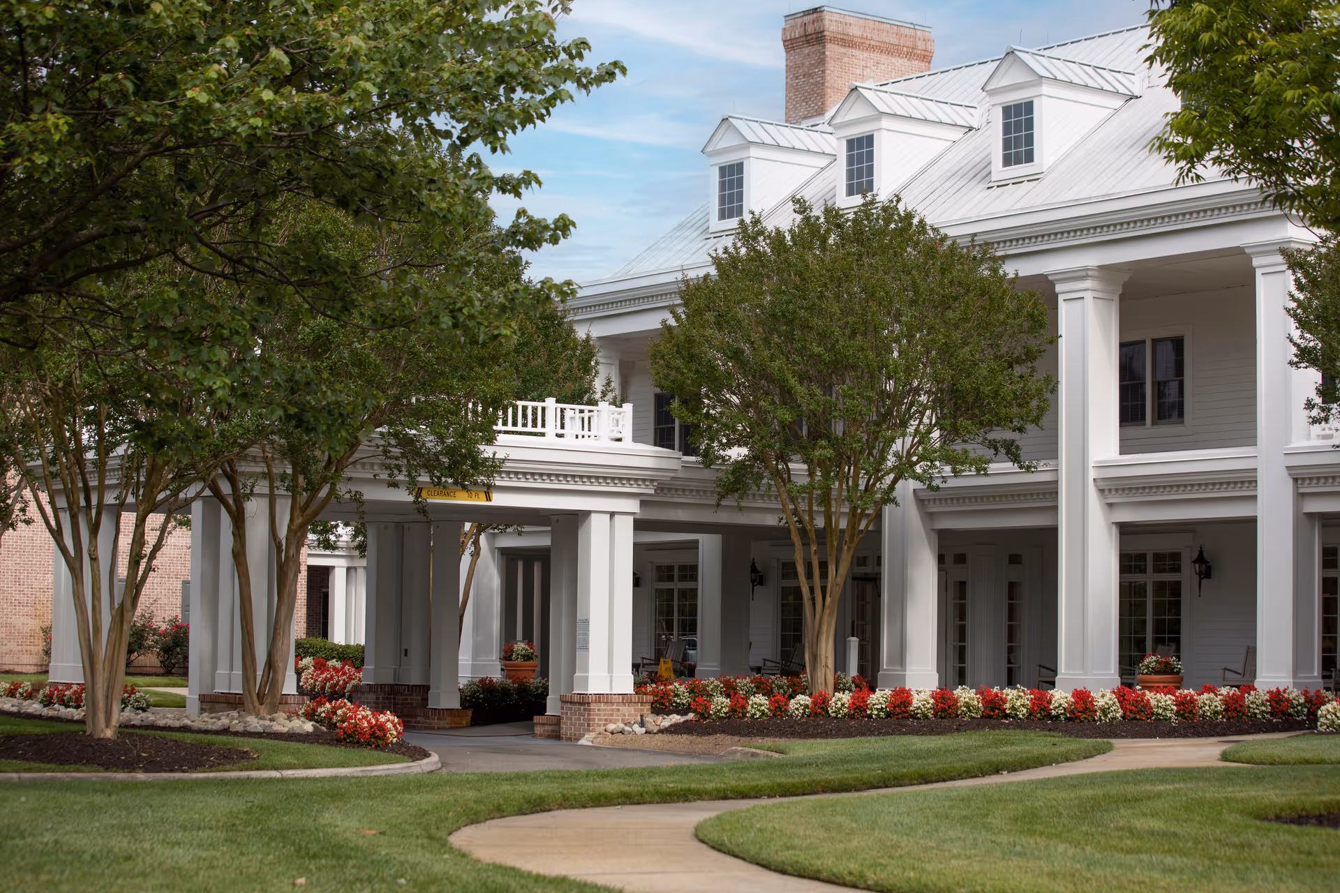 Exterior view of a white two-story building with a metal roof, large columns, and a covered entrance. The building is surrounded by well-maintained landscaping including green grass, trees, and flower beds with red and white flowers. A curved sidewalk leads to the entrance.