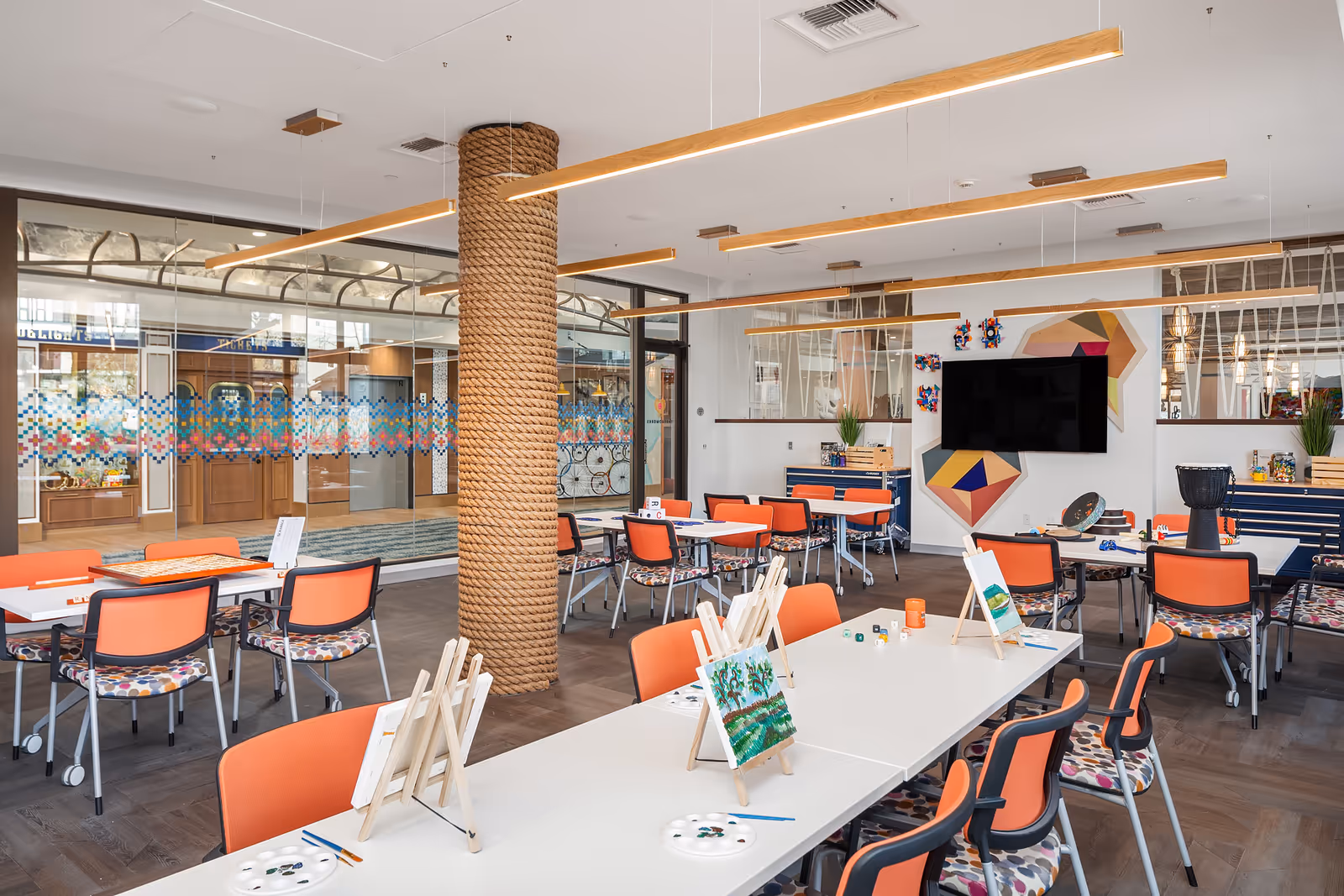 A bright and modern activity room with white tables and orange chairs featuring colorful patterned cushions. Several tables have small easels with paintings, paint palettes, and brushes, indicating an art activity setup. The room has a large flat-screen TV mounted on the wall, geometric wall art, and a column wrapped in thick rope. Large glass windows and doors reveal an adjacent area with wooden doors and a patterned glass partition.