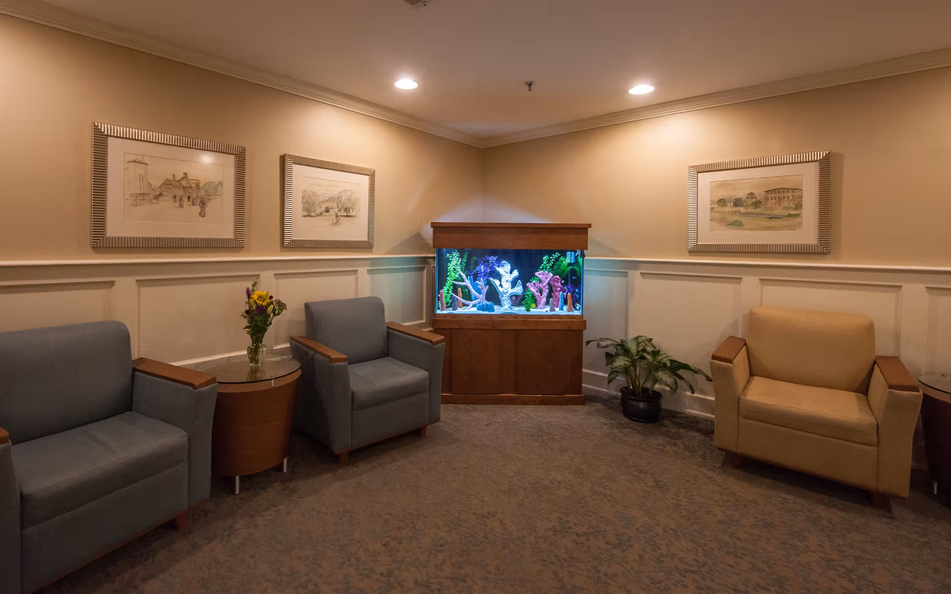 A cozy seating area in a senior living facility with two gray armchairs and one beige armchair arranged around a wooden fish tank filled with colorful coral decorations. There are two small round wooden side tables with glass tops, one holding a vase with flowers. The walls are beige with white wainscoting and decorated with three framed paintings. A potted plant is placed on the floor near the fish tank.