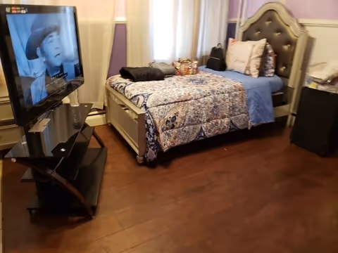 A bedroom with a bed covered in a patterned quilt and pillows, a flat-screen TV on a stand showing a black and white image of a man wearing a hat, purple walls, and hardwood flooring.