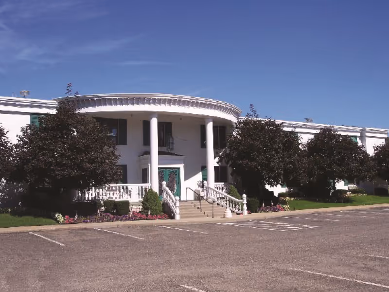 White two-story nursing facility with a columned portico, front steps and railing, trees and a parking lot in front.