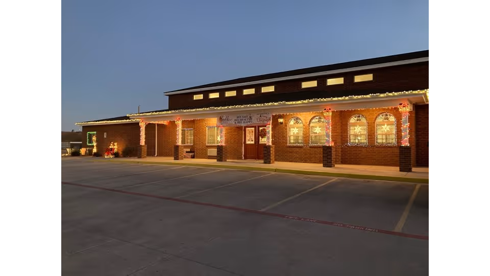 Exterior view of a brick building at dusk decorated with Christmas lights along the roofline and columns. The building has arched windows with illuminated star decorations inside. There is a banner above the entrance that reads 'BeeHive Homes Of Andrews' along with a holiday greeting. The parking lot in front is empty.