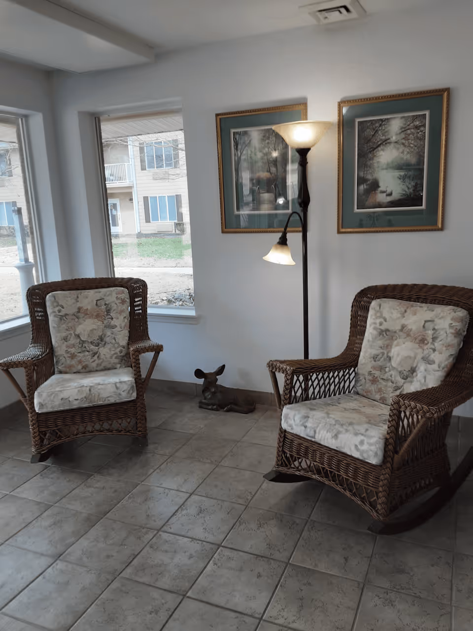 A cozy sitting area with two wicker rocking chairs featuring floral cushions, a floor lamp with two lights, and two framed nature-themed paintings on the wall. There is a small decorative deer figurine on the floor between the chairs. Large windows provide a view of the outside building and greenery.