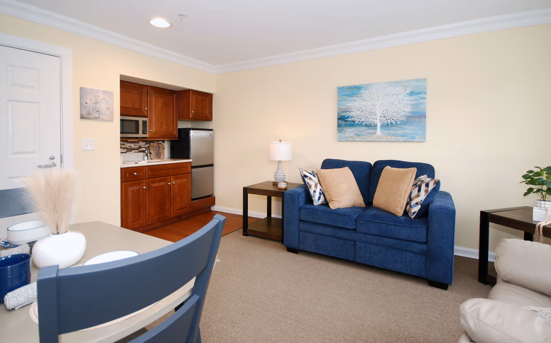 A cozy living area in a senior living facility featuring a blue loveseat with four decorative pillows, a side table with a lamp, and a painting of a white tree on a blue background hanging on the wall. To the left, there is a small kitchen area with wooden cabinets, a microwave, and a stainless steel refrigerator. In the foreground, part of a dining table with blue chairs and a white vase with dried pampas grass is visible.
