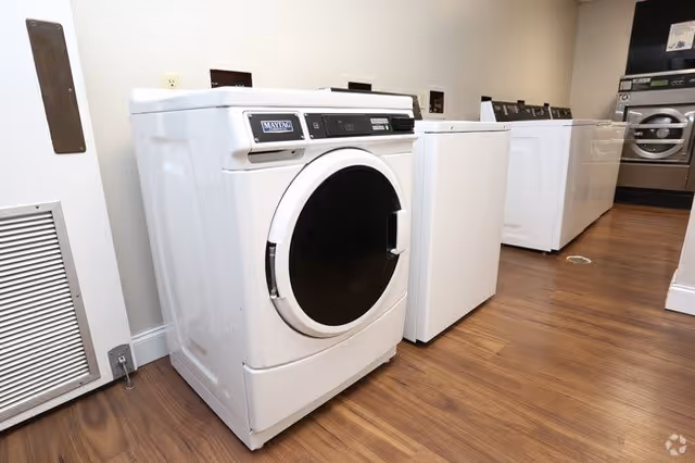 A row of white laundry machines including washers and dryers in a laundry room with wooden flooring and beige walls.