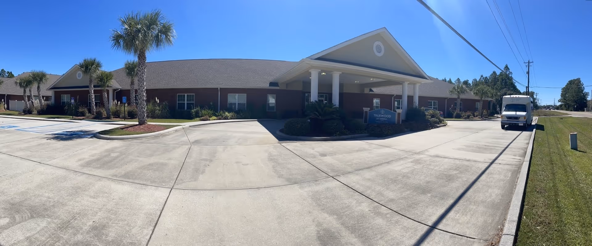 Exterior view of a single-story brick building with a covered entrance supported by white columns. There are palm trees and landscaped bushes around the building. A white van is parked on the driveway to the right. The sky is clear and blue.