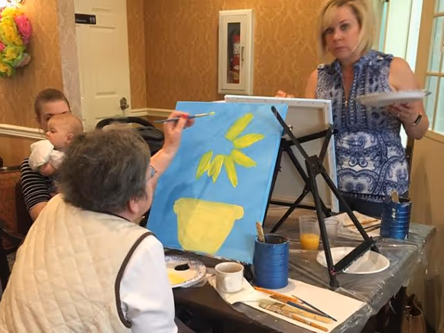 An elderly woman painting a yellow flower and pot on a blue canvas at an easel, with a younger woman standing nearby holding a palette. In the background, another woman is holding a baby while seated. The setting appears to be a cozy indoor room with patterned wallpaper and a fire extinguisher mounted on the wall.