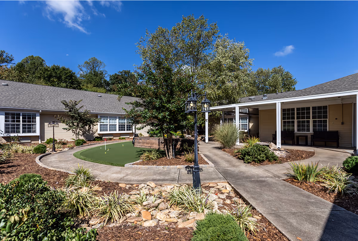 Outdoor courtyard area at Richland Square featuring a small putting green, paved walkways, landscaped garden beds with shrubs and plants, a black lamppost, and beige buildings with white-framed windows under a clear blue sky.