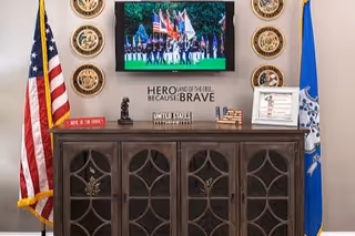 A patriotic display featuring an American flag on the left and a Connecticut state flag on the right, with a dark wooden cabinet below. On the wall above the cabinet is a mounted flat-screen TV showing a color guard with multiple flags. Surrounding the TV are six military emblems. Below the TV is a sign that reads 'HERO AND OF THE FREE BECAUSE BRAVE' along with other patriotic decorations including a small bust, a framed certificate, and a small American flag.